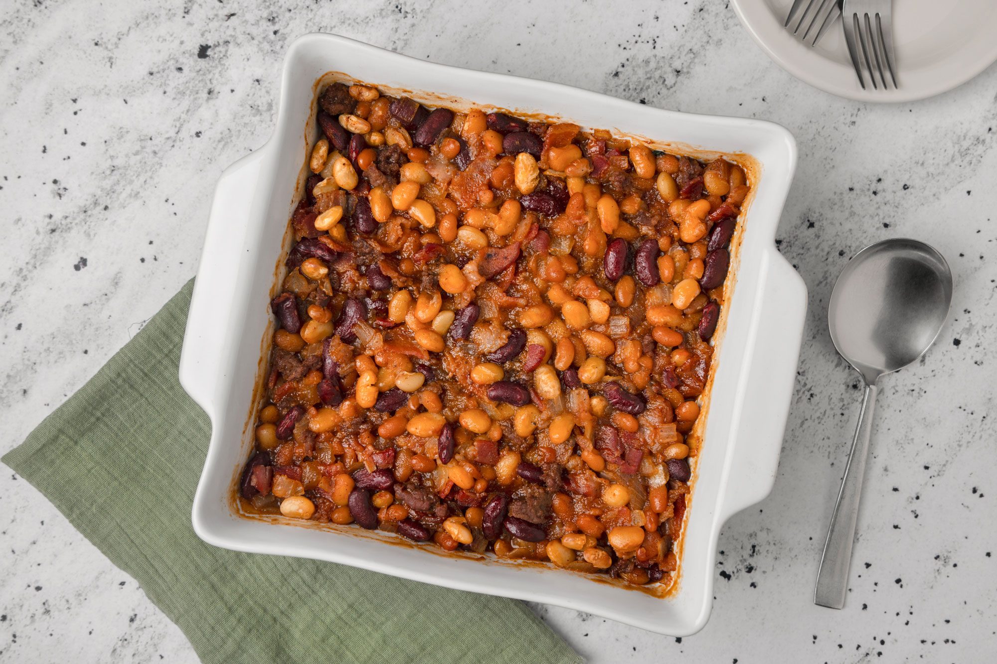 Top Shot of a square white baking dish of baked beans in tomato sauce rests on a green cloth; a metal spoon and two stacked plates are beside it on a speckled countertop; creating a cozy meal setting