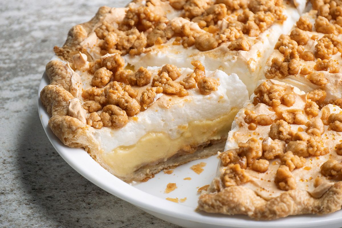 A close-up of a creamy pie with a crumbly topping and a golden crust, served in a white pie dish. One slice has been removed, revealing the smooth, layered filling inside.