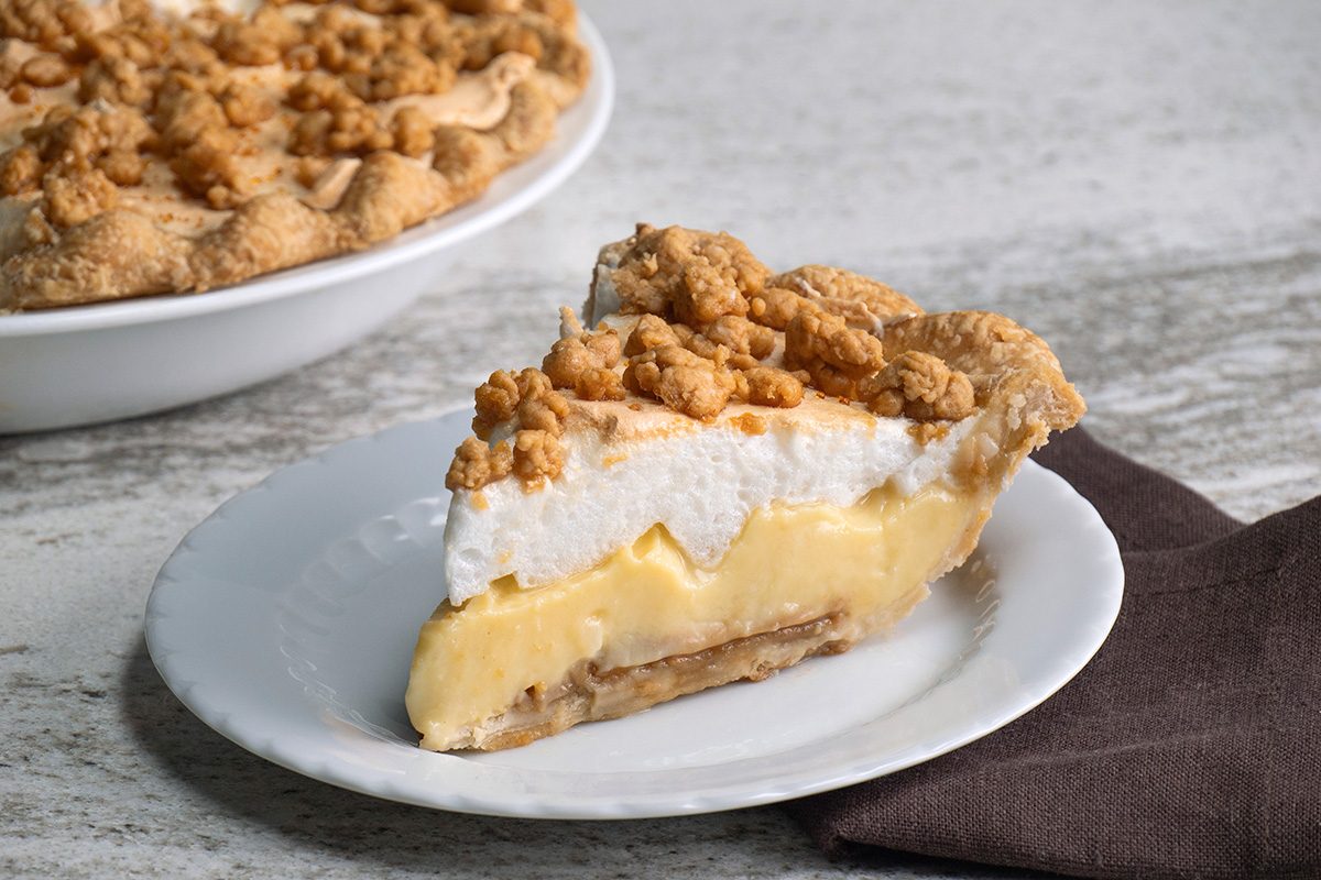 A slice of pie with a crumbly topping, meringue layer, and creamy yellow filling sits on a white plate, with the rest of the pie in the background on a gray surface next to a brown napkin.