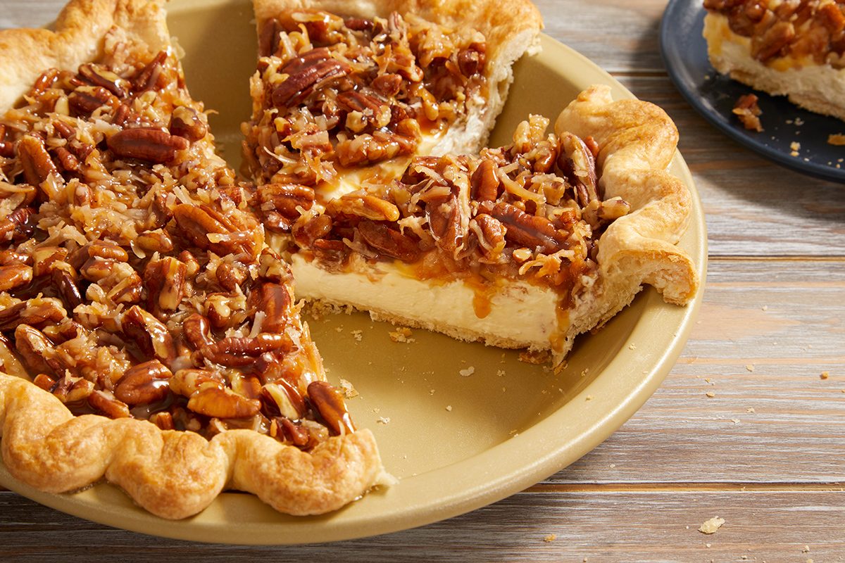 A pecan pie with a golden crust and creamy filling sits on a wooden table. One slice is missing from the pie, and the slice is placed on a separate blue plate in the background.