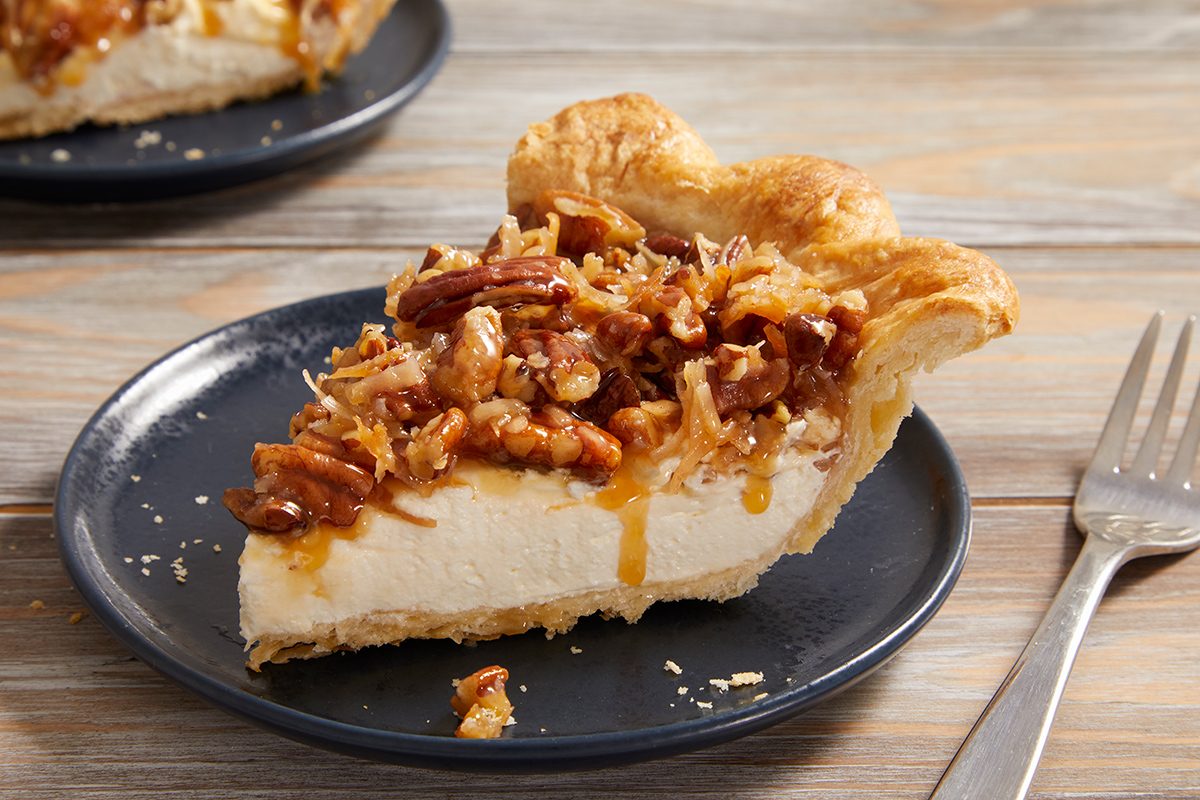A slice of creamy pie topped with caramel, pecans, and toasted coconut sits on a dark plate, with a fork beside it on a wooden table. Another plate with pie is in the background.