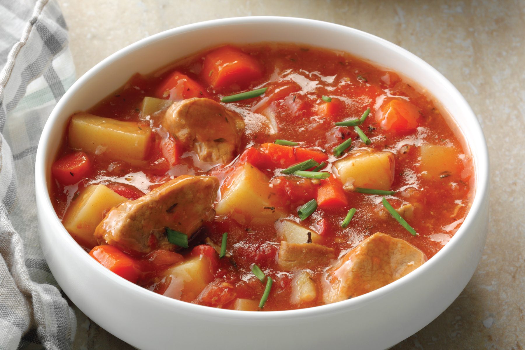 A white bowl filled with a chunky pork vegetable stew containing pieces of meat, potatoes, red bell peppers, and garnished with chopped fresh herbs. A striped cloth napkin is beside the bowl.