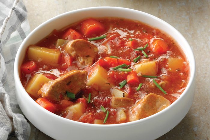 A white bowl filled with a chunky pork vegetable stew containing pieces of meat, potatoes, red bell peppers, and garnished with chopped fresh herbs. A striped cloth napkin is beside the bowl.