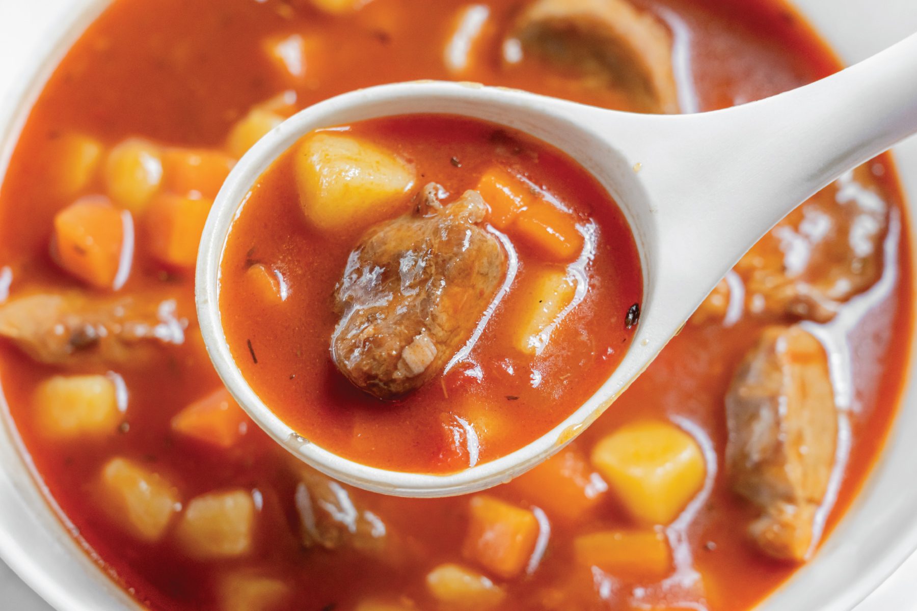 A close-up of a white spoon holding a serving of pork vegetable soup with chunks of beef, potatoes, and carrots in a rich, red broth. The bowl below shows more of the same stew.