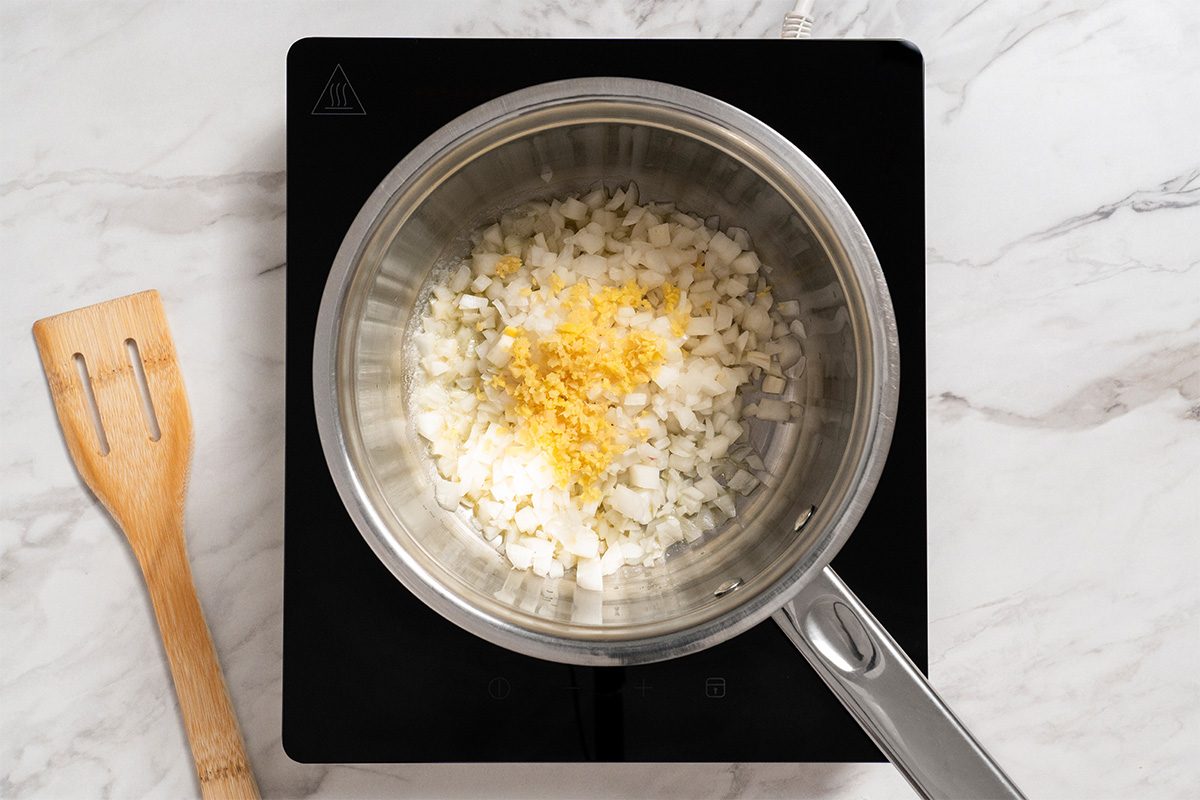 A saucepan on a stovetop contains chopped onions and grated lemon zest. A wooden spatula rests beside the stove on a white marble countertop.