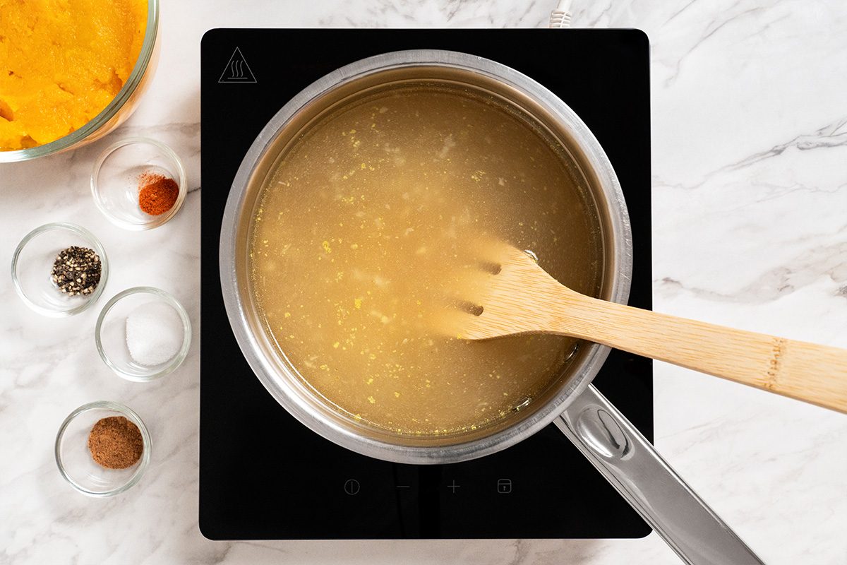 A pot of broth is simmering on a black stovetop, being stirred with a wooden spoon. Small bowls of spices and a bowl with a yellow substance are arranged nearby on a marble countertop.