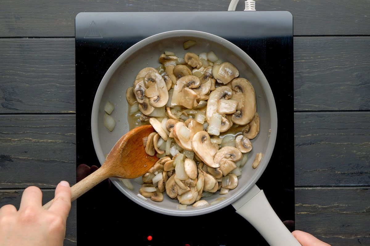 Top shot of A hand stirs sliced mushrooms and chopped onions with a wooden spoon in a white frying pan on an electric stovetop; kitchen background is visible; cooking process appears active
