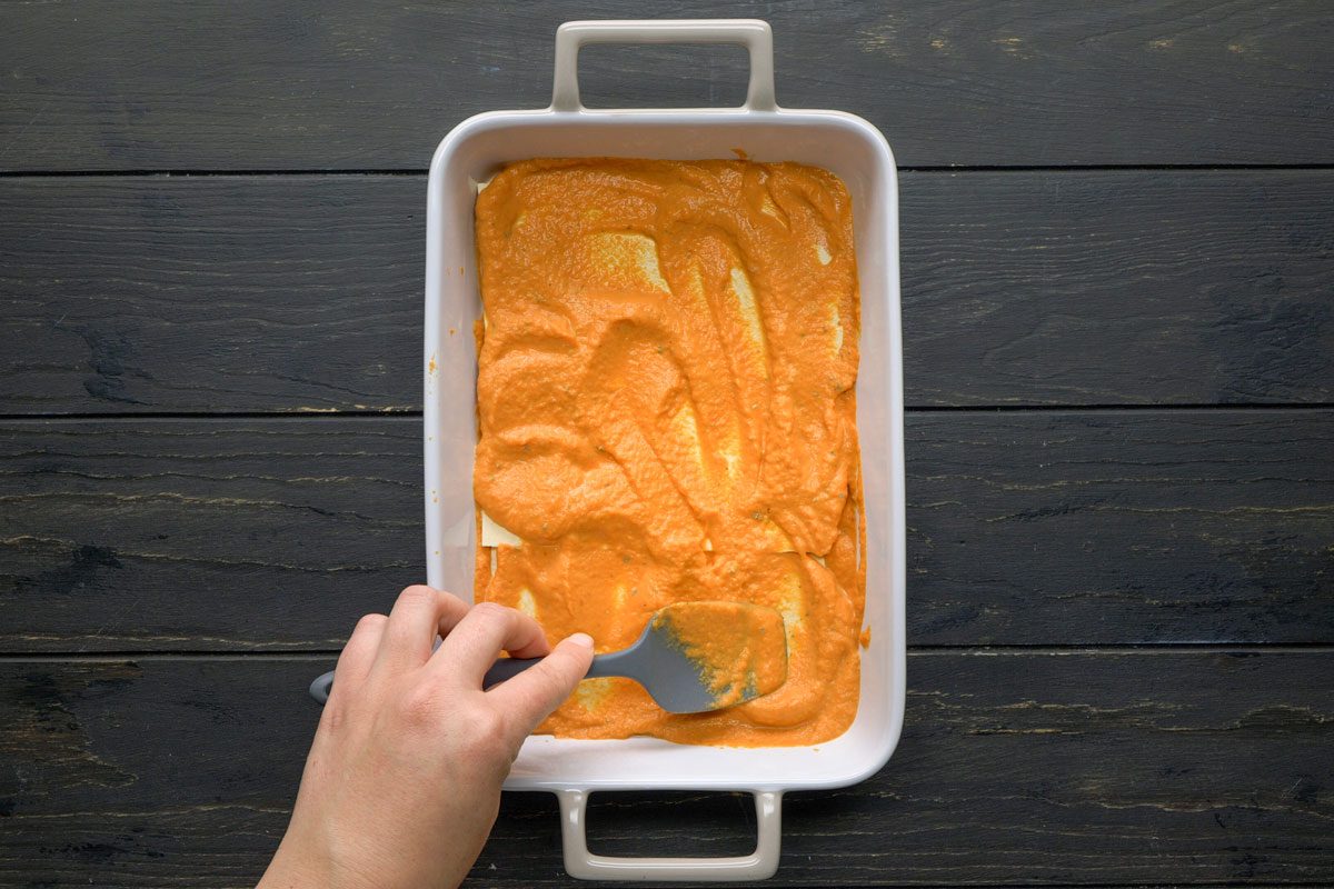 Top Shot of A hand spreads orange sauce with a spatula in a rectangular white baking dish; the background features a dark wooden surface, creating a warm and rustic kitchen atmosphere