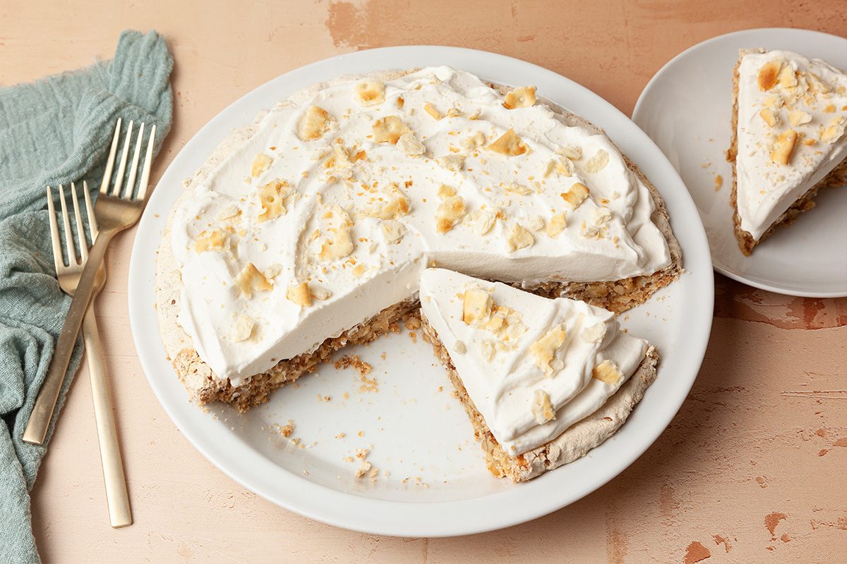 A cream pie with a crumbly crust sits on a white plate, topped with whipped cream and cookie crumbles. One slice is missing and placed on a small plate nearby. Two forks rest beside a blue napkin.
