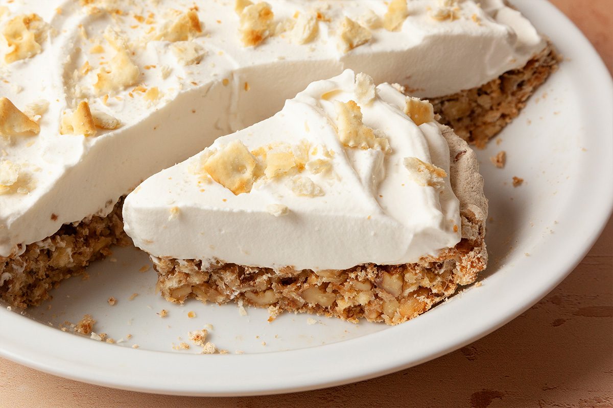A slice of creamy pie with a nutty, crumbly crust is served on a white plate, with whipped topping and crushed nuts sprinkled on top.