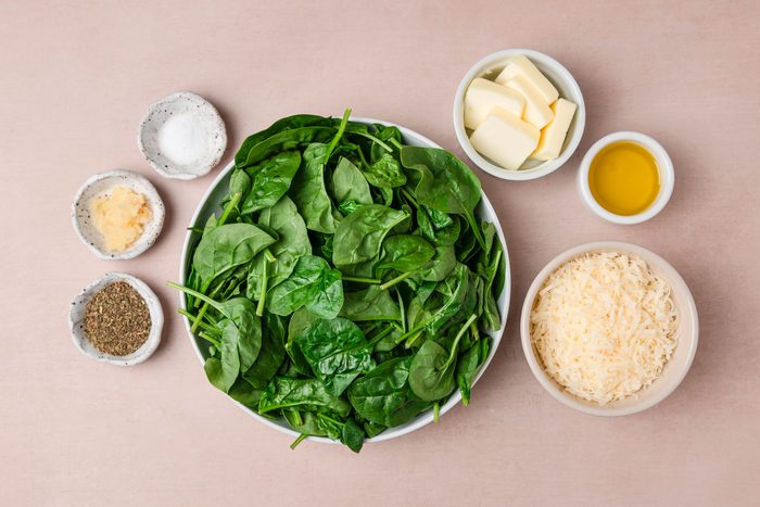 A top shot of a bowl of vibrant spinach is surrounded by small bowls containing butter; olive oil; shredded cheese; dried herbs; minced garlic; and salt on a light brown surface, creating a fresh and inviting meal preparation scene