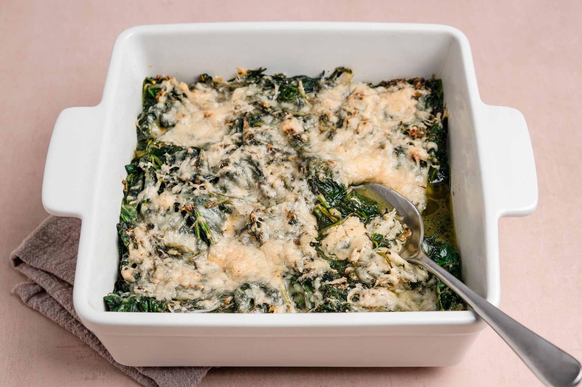 A 3/4th shot of a white square baking dish of cheesy baked spinach; melted cheese topping; set on a beige surface; accompanied by a metal serving spoon and gray cloth underneath