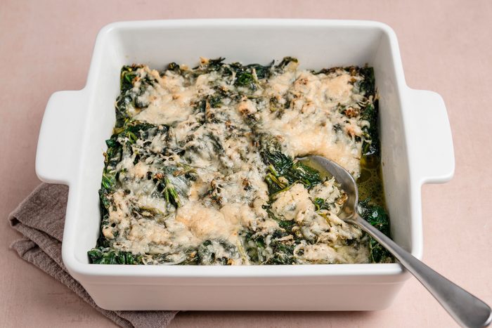 A 3/4th shot of a white square baking dish of cheesy baked spinach; melted cheese topping; set on a beige surface; accompanied by a metal serving spoon and gray cloth underneath