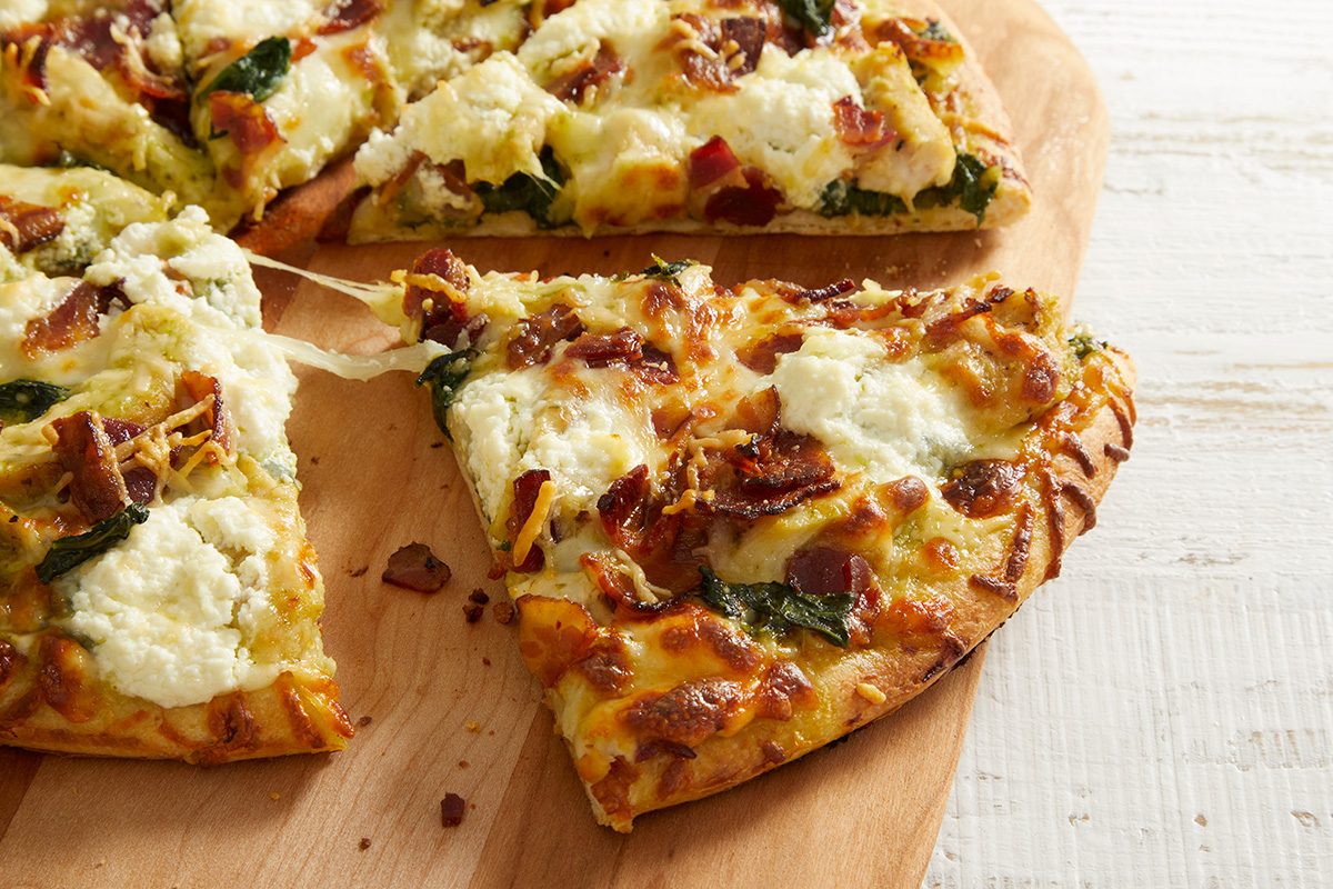 A close-up of a sliced pizza on a wooden board. The pizza has a golden crust, melted cheese, spinach, chunks of bacon, and dollops of ricotta cheese. One slice is slightly pulled away from the rest.