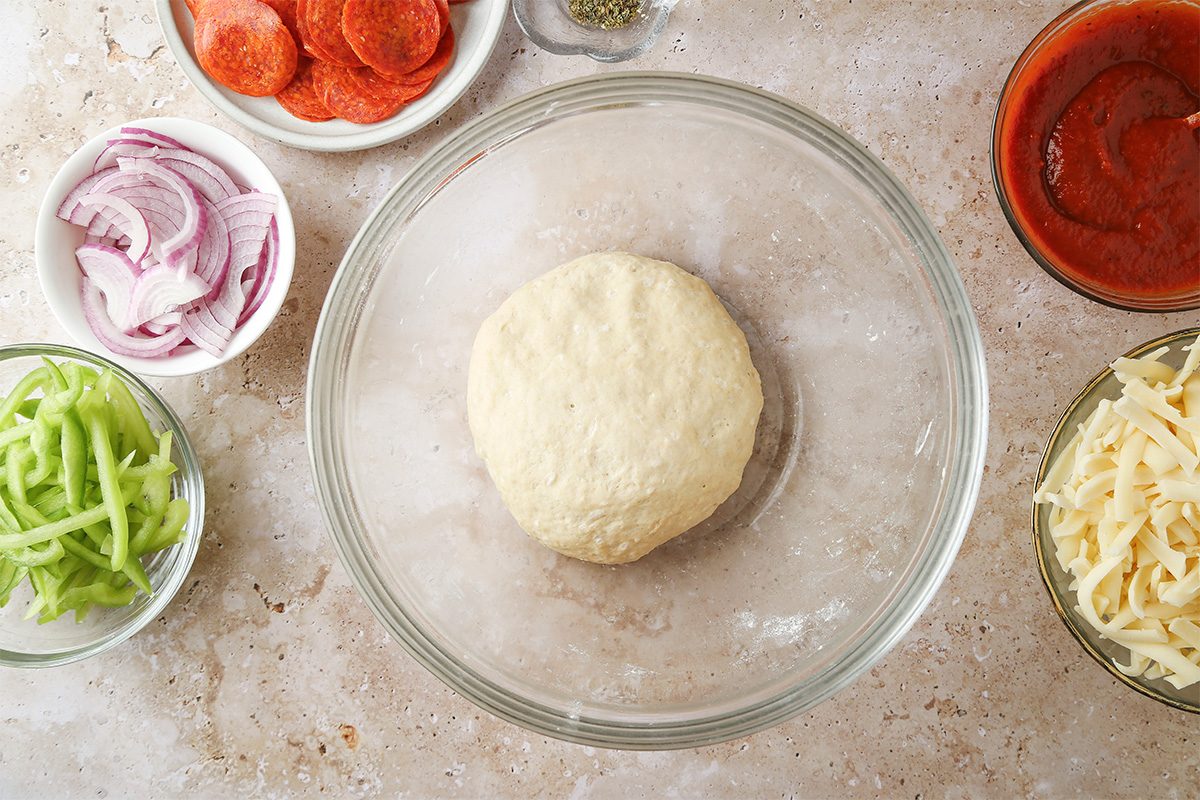 A ball of pizza dough in a glass bowl surrounded by bowls of sliced pepperoni, red onions, green bell peppers, shredded cheese, pizza sauce, and dried herbs on a countertop.
