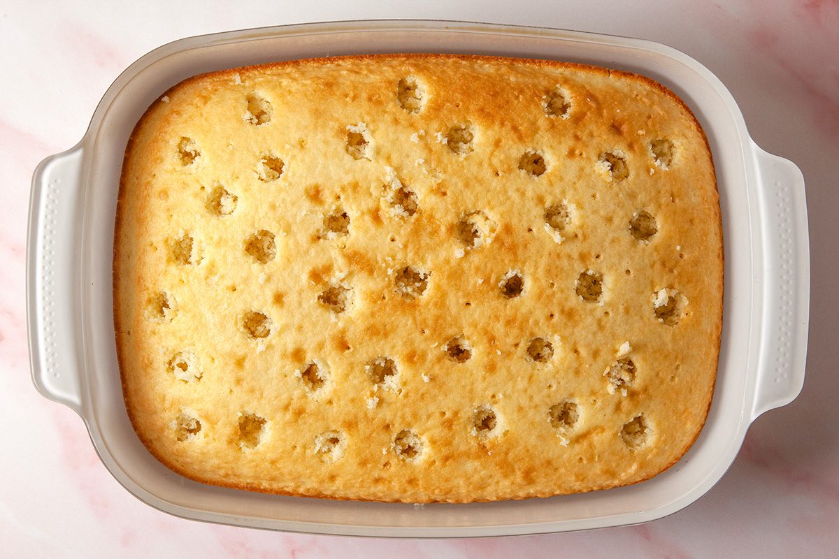 overhead shot of a rectangular baked cake in a white dish with evenly spaced holes poked across the surface, viewed from above on a light pink marble background