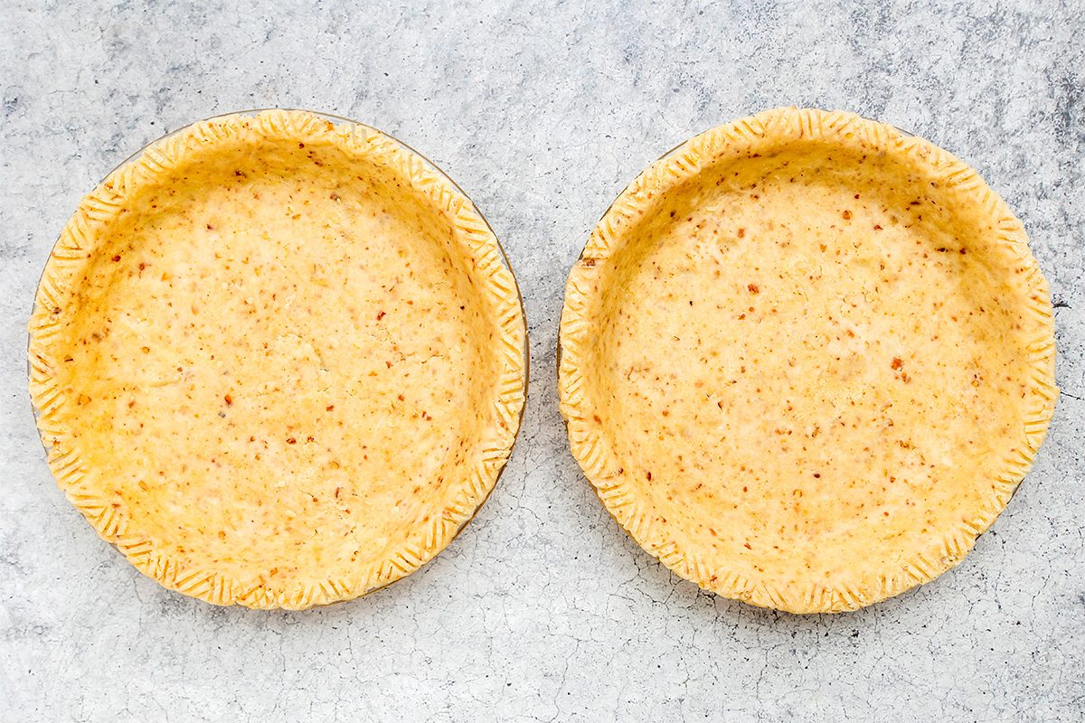 Two empty, unbaked pie crusts in round pans sit side by side on a light, textured surface. The edges of each crust are neatly crimped with a fork pattern.