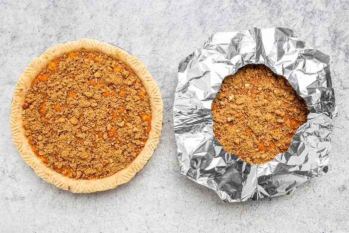 Two unbaked crumb-topped pies on a gray surface; one pie has exposed edges, while the other has its crust edges covered with crinkled aluminum foil.