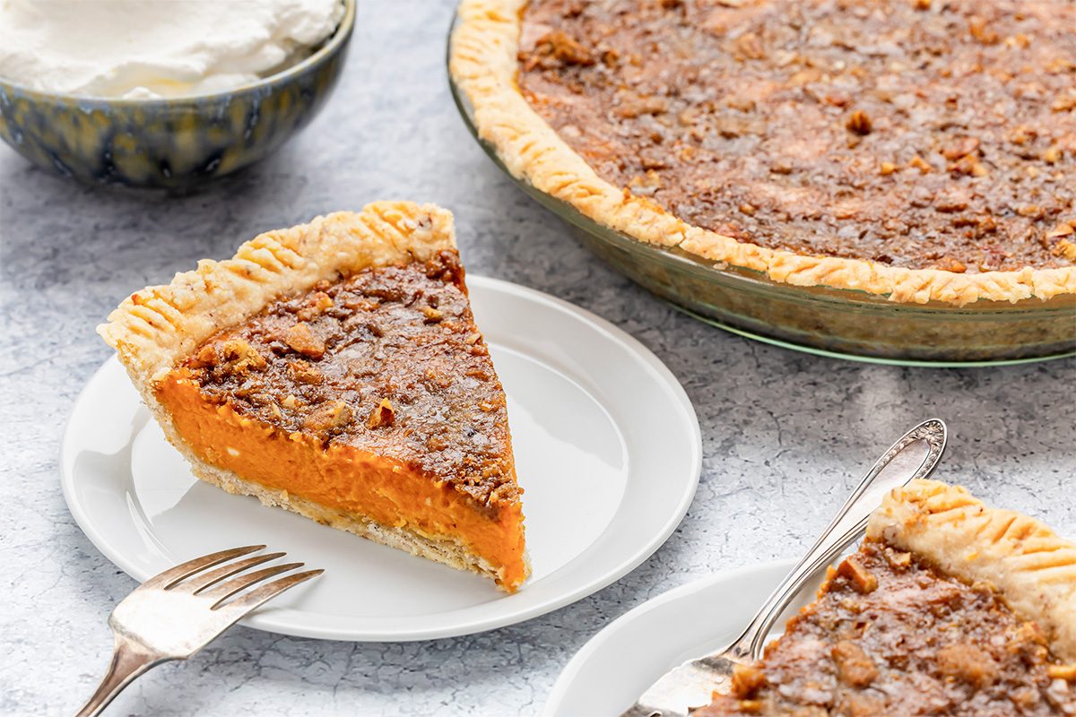 A slice of pecan pie on a white plate with a fork, a whole pecan pie in the background, and a bowl of whipped cream beside them on a light textured surface.