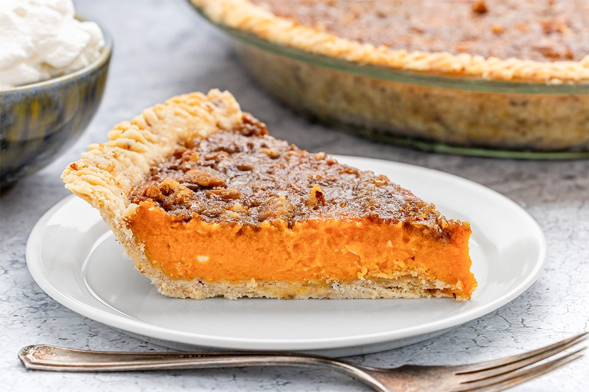 A slice of sweet potato pie with a crumbly topping sits on a white plate, with a fork beside it. In the background, there is a pie dish and a bowl of whipped cream.