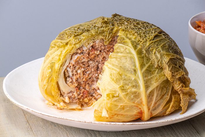 A stuffed cabbage head sits on a white plate, with a wedge cut out to reveal a filling of rice and ground meat. The cabbage leaves are cooked and slightly wilted, with the filling clearly visible inside.