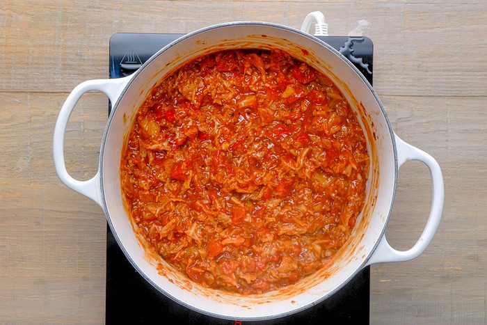 A white pot filled with thick, chunky red tomato sauce simmers on a black stovetop, with visible bits of vegetables and herbs throughout.