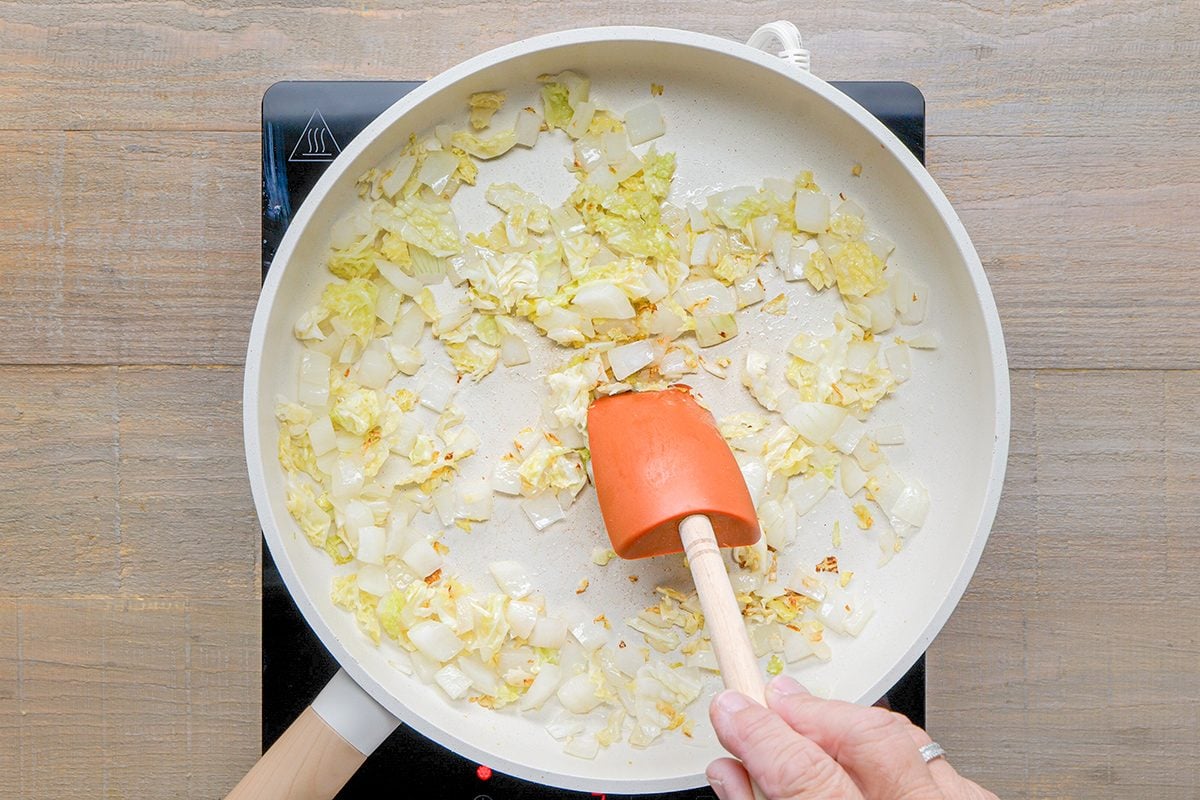 A hand stirs chopped onions cooking in a white frying pan with an orange spatula on a stovetop.