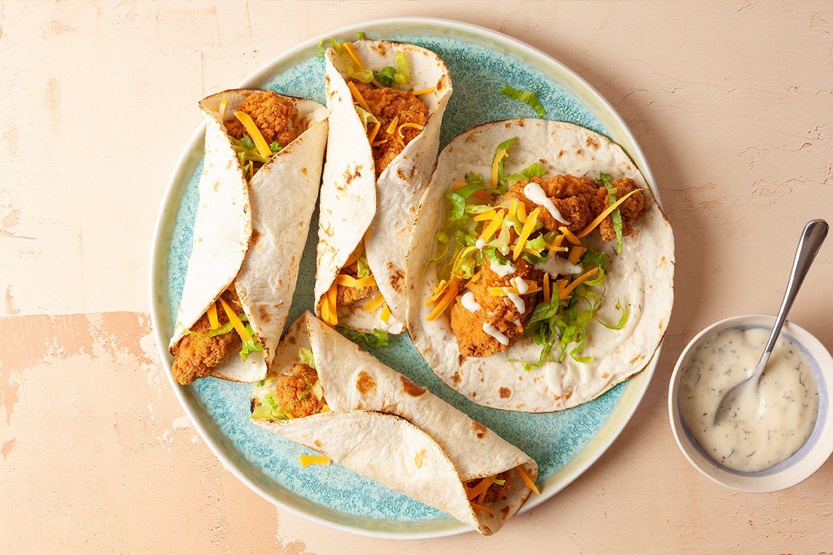 A plate with four soft tacos filled with crispy breaded chicken, shredded lettuce, and cheese, next to a small bowl of creamy sauce with a spoon on a light beige surface.