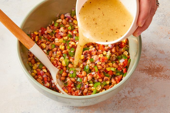 A hand pours a bowl of yellow vinaigrette over a colorful bean and vegetable salad in a large mixing bowl with a wooden spoon.