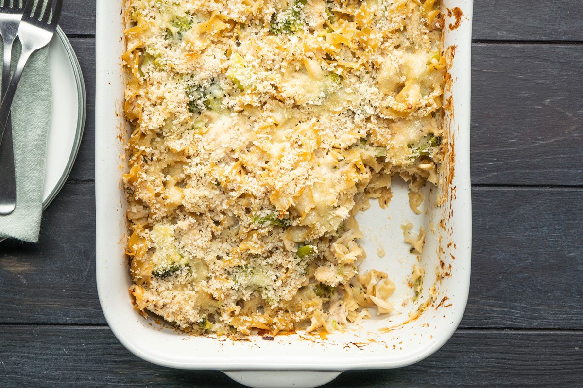 Top Shot of a golden-brown baked casserole rests in a white dish; partly served, revealing its layers; surrounded by plates; forks; and a napkin on a dark wooden table. The scene suggests comfort food and communal dining