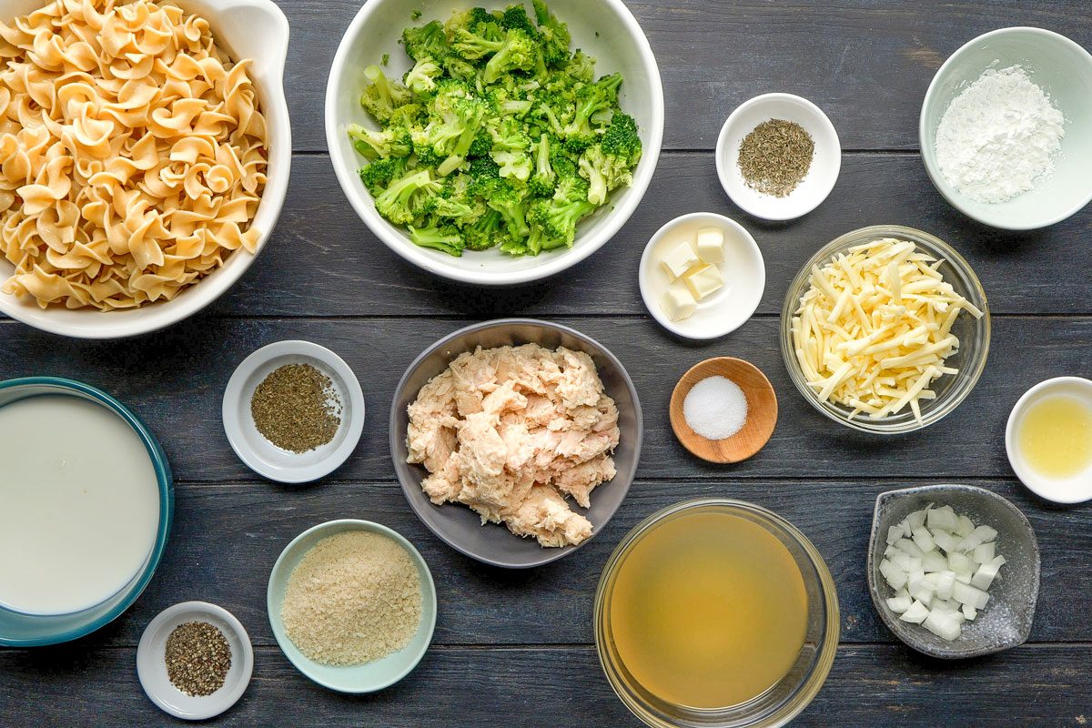 A top shot shows bowls with egg noodles; broccoli; shredded cheese; cooked chicken; milk; seasoning; onions; broth; flour and spices on a dark background; Tuna Broccoli Casserole
