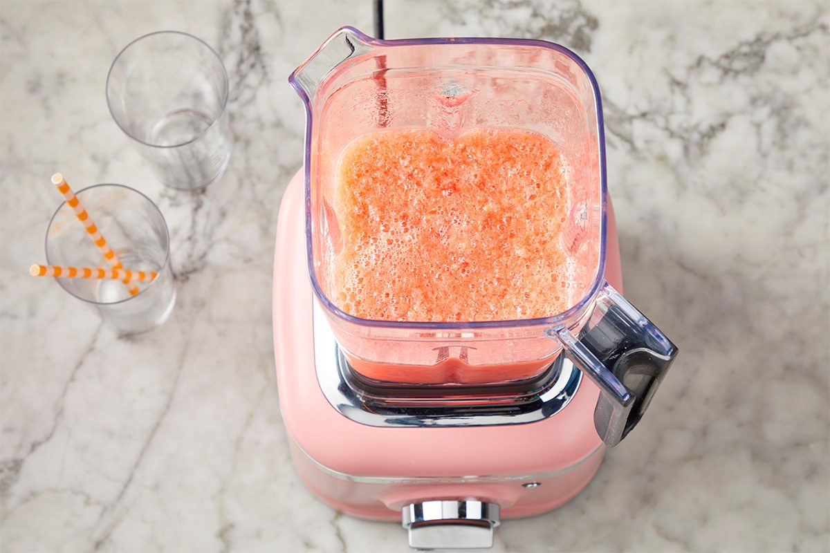 A pink blender filled with a frothy orange smoothie sits on a marble countertop next to two empty glasses with orange striped straws.