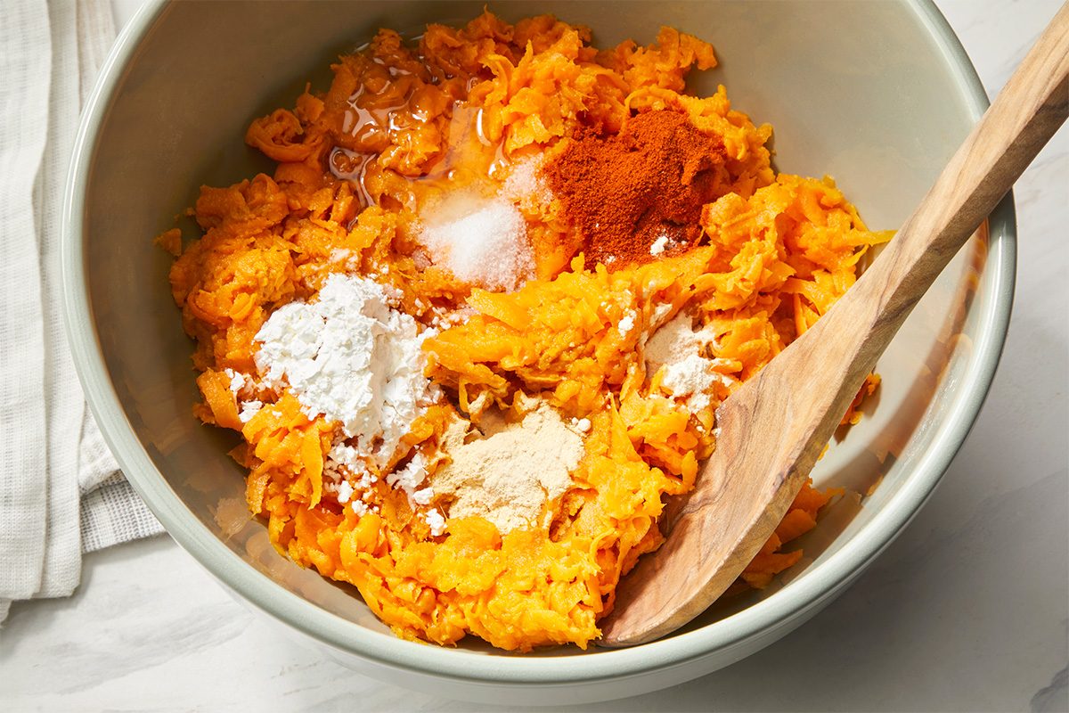 A mixing bowl filled with shredded sweet potatoes, white powder (likely flour or starch), spices, and a wooden spoon resting inside, ready for mixing. A folded white cloth sits nearby on the counter.