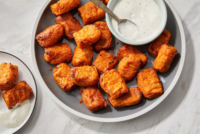 A gray plate filled with crispy, golden-brown tater tots is shown, with a small bowl of creamy white dipping sauce and a spoon. A separate small plate holds more tots with a dollop of sauce.