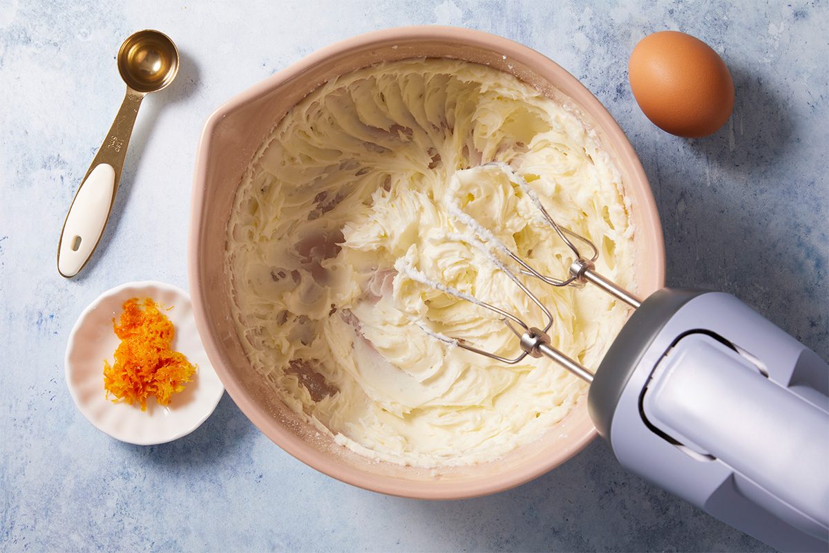 A mixing bowl with creamy batter and a hand mixer, next to an egg, a measuring spoon, and a small dish containing orange zest, all on a light blue surface.