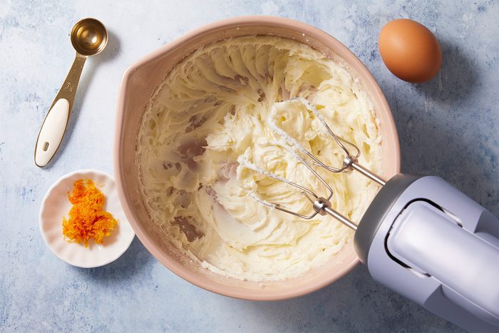 A mixing bowl with creamy batter and a hand mixer, next to an egg, a measuring spoon, and a small dish containing orange zest, all on a light blue surface.