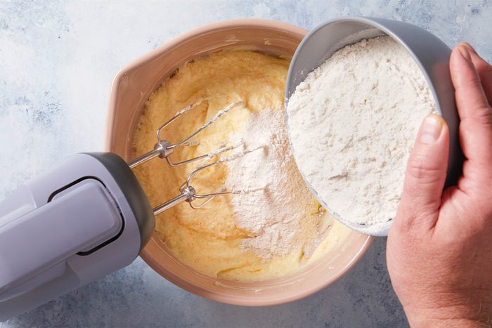 A person pours flour from a bowl into a mixing bowl with batter while using an electric hand mixer, preparing ingredients for baking.