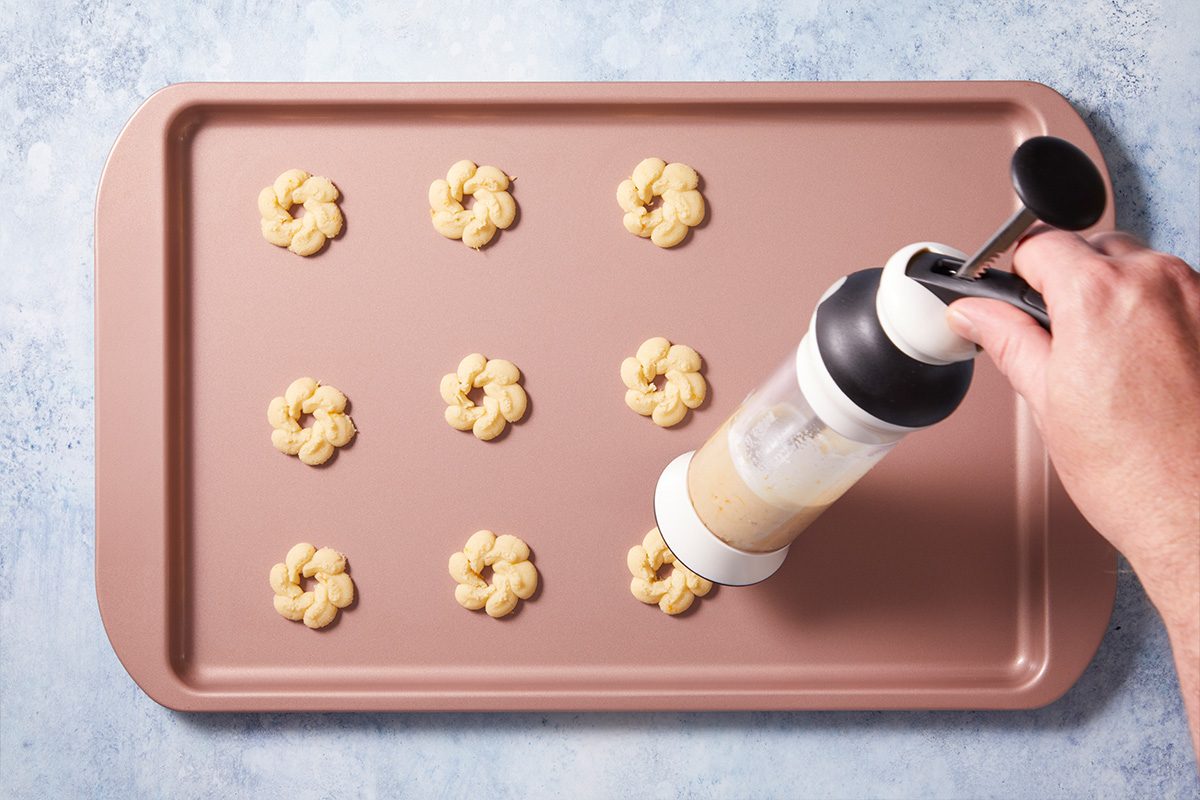A hand holds a cookie press, piping flower-shaped cookie dough pieces onto a pink baking sheet arranged in three rows on a light blue surface.