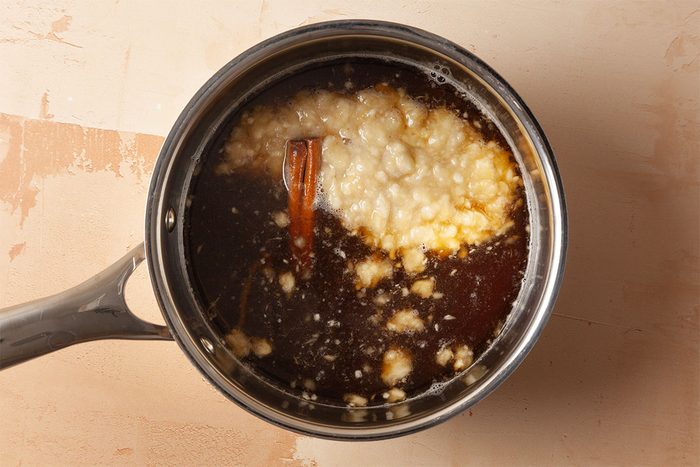 A saucepan filled with a brown liquid, chunks of butter, and a cinnamon stick, sitting on a light-colored surface.
