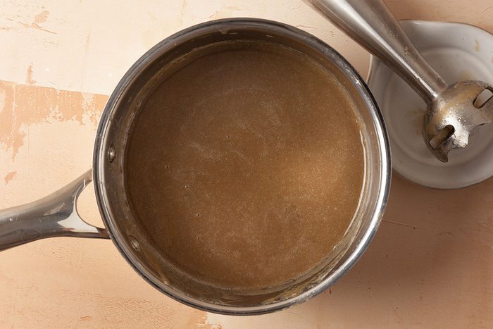 A saucepan filled with light brown gravy sits on a light-colored surface. Next to it is an immersion blender resting on a small dish.