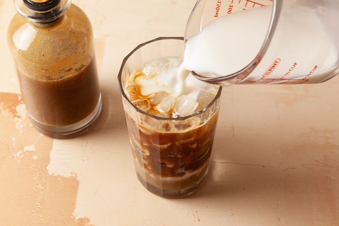 A glass of iced coffee is being topped with milk from a measuring cup. A bottle of coffee concentrate sits nearby on a beige surface.