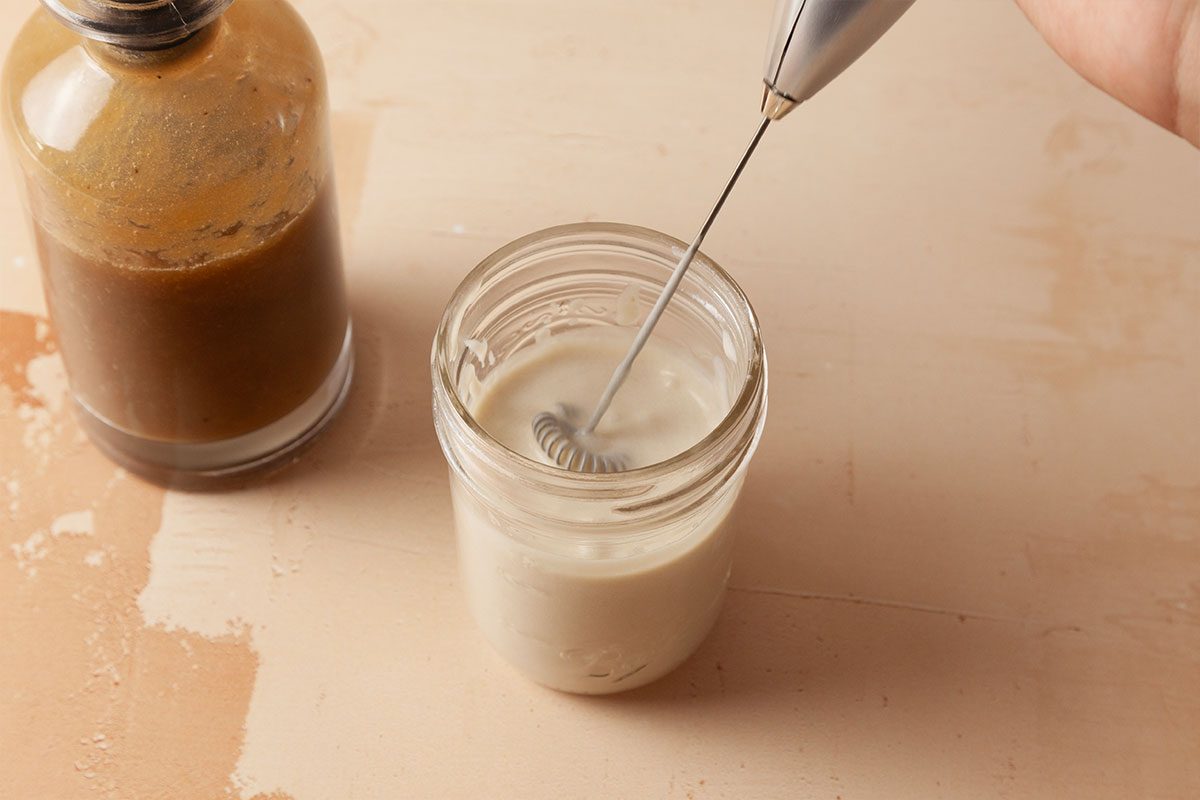 A hand uses a small electric whisk to froth milk in a glass jar, with a bottle of brown liquid, possibly coffee, placed nearby on a beige surface.