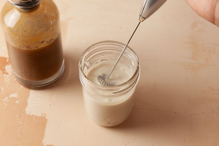 A hand uses a small electric whisk to froth milk in a glass jar, with a bottle of brown liquid, possibly coffee, placed nearby on a beige surface.
