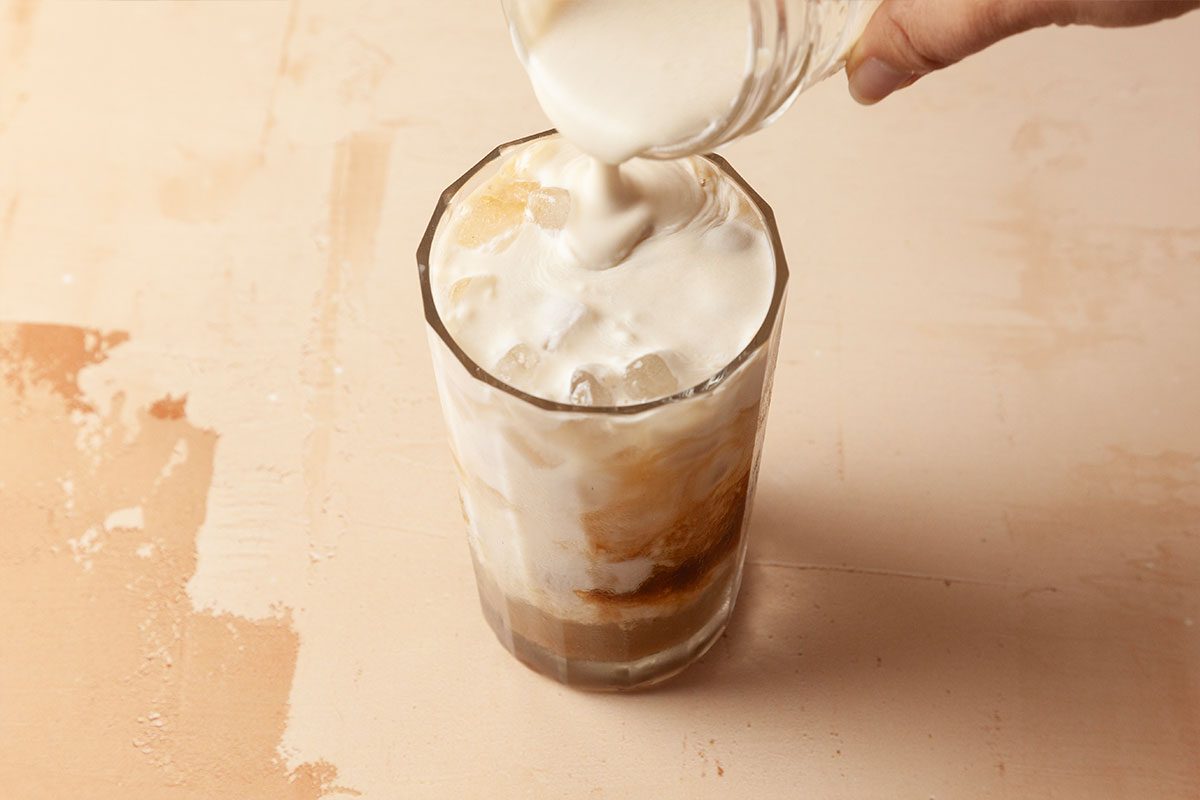 A hand pours creamy milk over a glass of iced coffee, creating swirls as the liquid mixes over the ice.