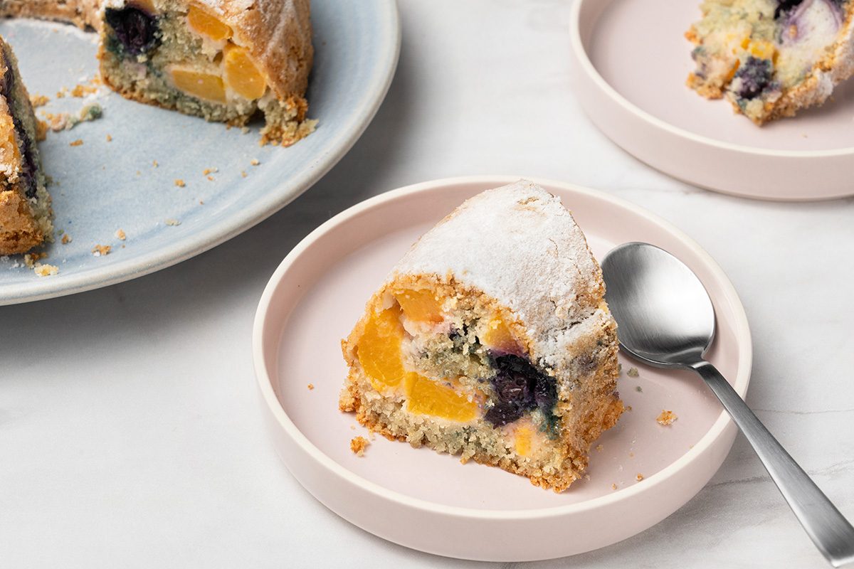 A slice of cake with visible pieces of peach and blueberry sits on a pale pink plate with a spoon. More cake is on a separate plate in the background. The cake is dusted with powdered sugar.