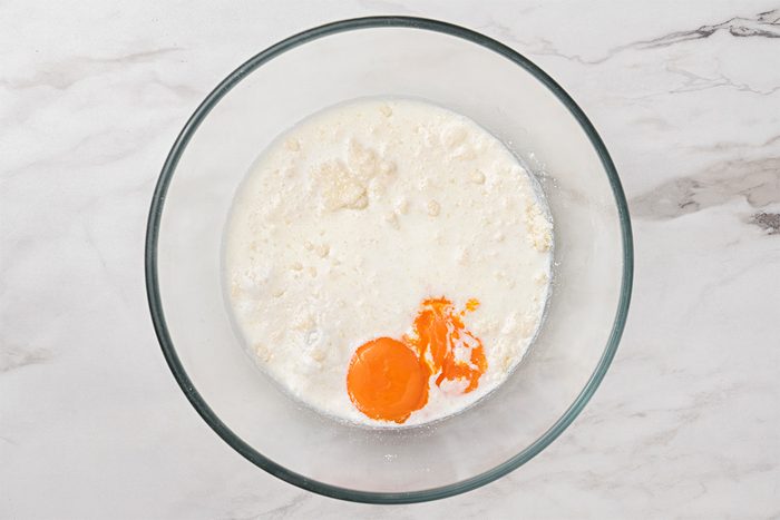 A glass bowl on a marble surface contains milk, flour, and two cracked eggs with visible yolks, ready to be mixed together.