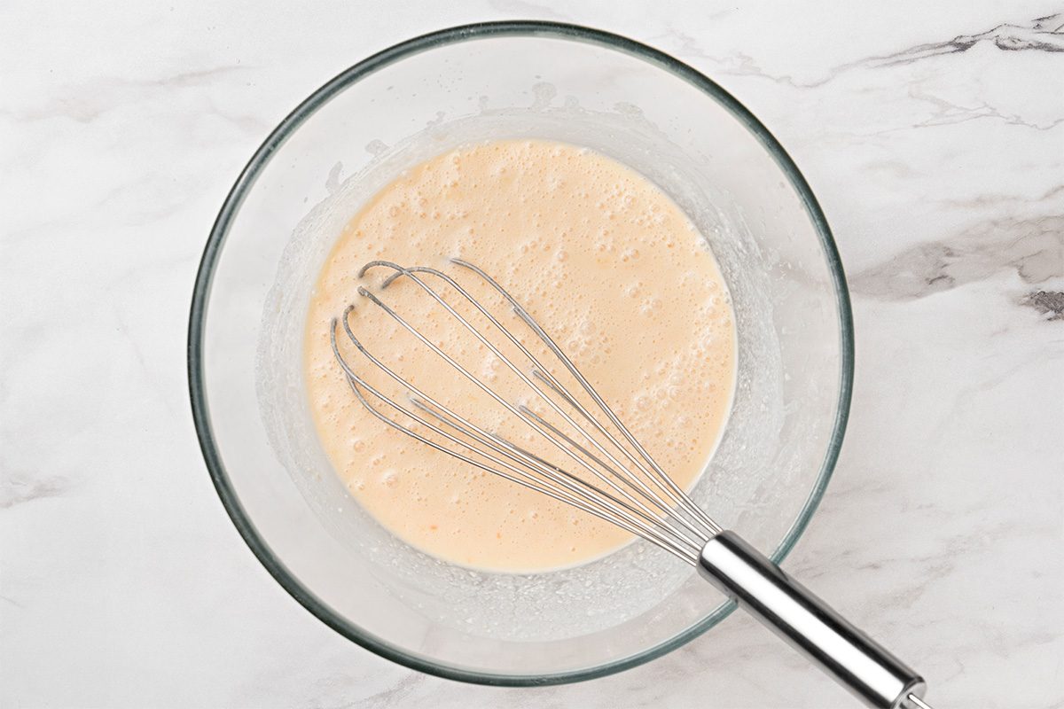 A glass mixing bowl with a light beige batter and a metal whisk on a white marble surface.