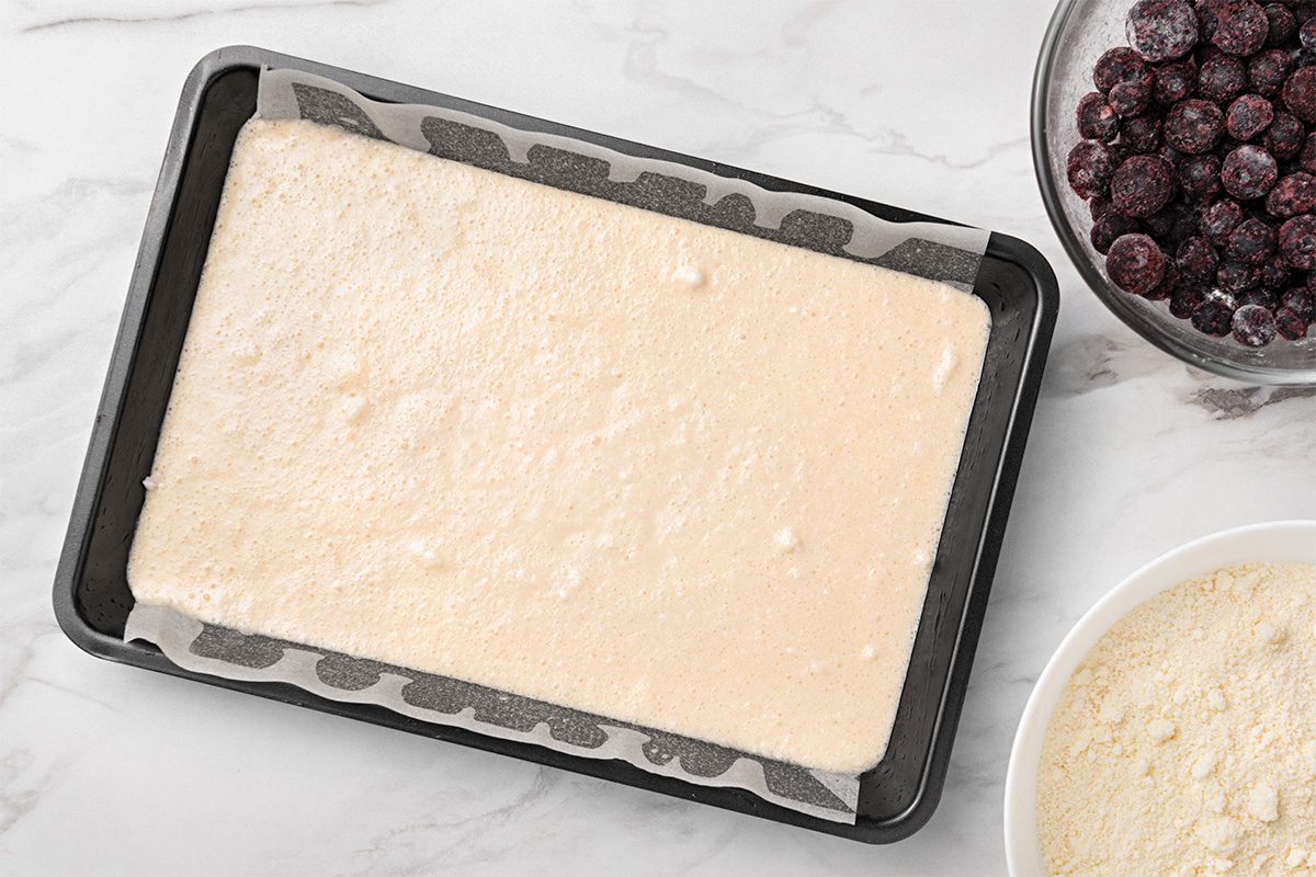 A rectangular baking tray lined with parchment paper holds a layer of pale batter. Nearby, there is a bowl of dark frozen berries and a bowl of light-colored flour on a white marble surface.