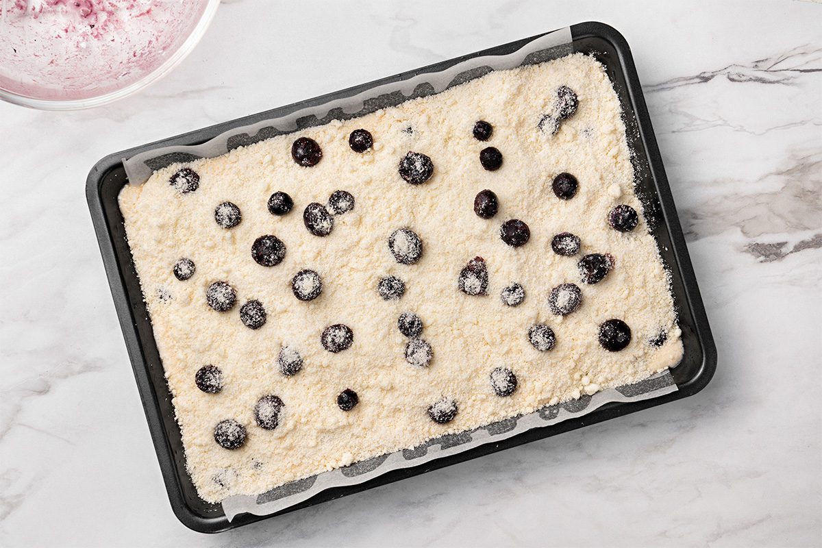 A rectangular baking pan lined with parchment paper holds an unbaked cake batter topped with scattered blueberries on a marble countertop.
