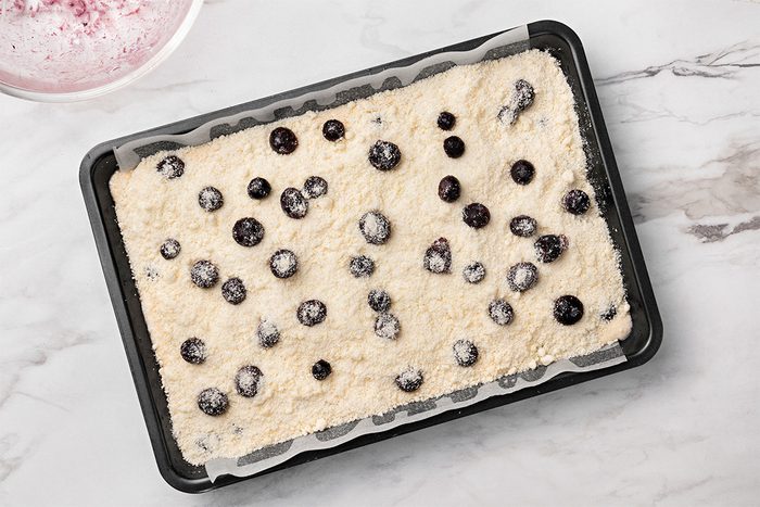 A rectangular baking pan lined with parchment paper holds an unbaked cake batter topped with scattered blueberries on a marble countertop.